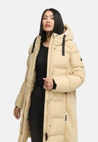 Beige lange pufferjas met een nepbont kraag, ritssluiting en zwarte bandjes. Voorzien van gestikte delen en zijzakken.