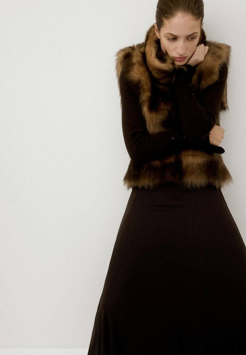 Chal de pelo sintético marrón sobre una blusa negra de manga larga ajustada, combinado con una falda negra fluida. Textura suave, forma elegante y diseño en capas.