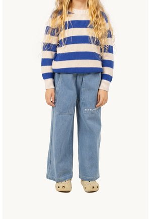 Enfant portant un pull rayé bleu et blanc, un jean bleu clair ample avec un texte sur la poche, et des chaussures beige sans lacets, debout devant un fond uni.