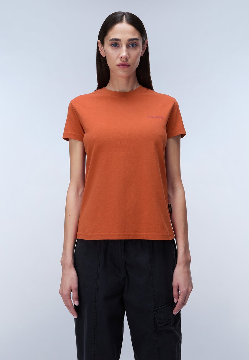 Camiseta de algodón naranja con mangas cortas y cuello redondo, que presenta un pequeño logo púrpura en el pecho. Llevada con pantalones cargo negros.