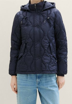 Person iført en marineblå quiltet hættetrøje med knap og lynlås, kombineret med lyseblå jeans.