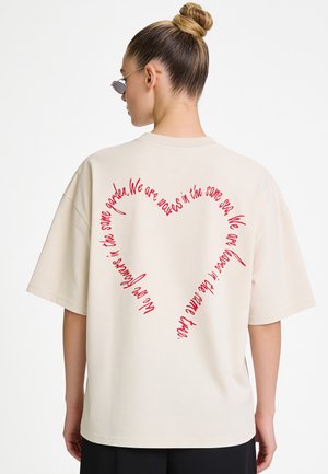 Femme avec les cheveux en chignon, portant des lunettes de soleil et un t-shirt crème oversize avec un texte rouge formant un cœur dans le dos.