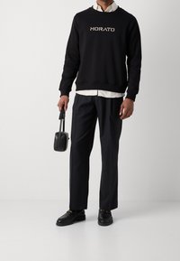 Sweat-shirt noir avec le logo "MORATO", chemise blanche à col en dessous, pantalon noir et chaussures noires. Tenant un petit sac noir.