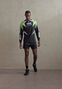 Man walking indoors wearing black and green long-sleeve sports shirt, black shorts, black socks, and black sneakers against plain wall.