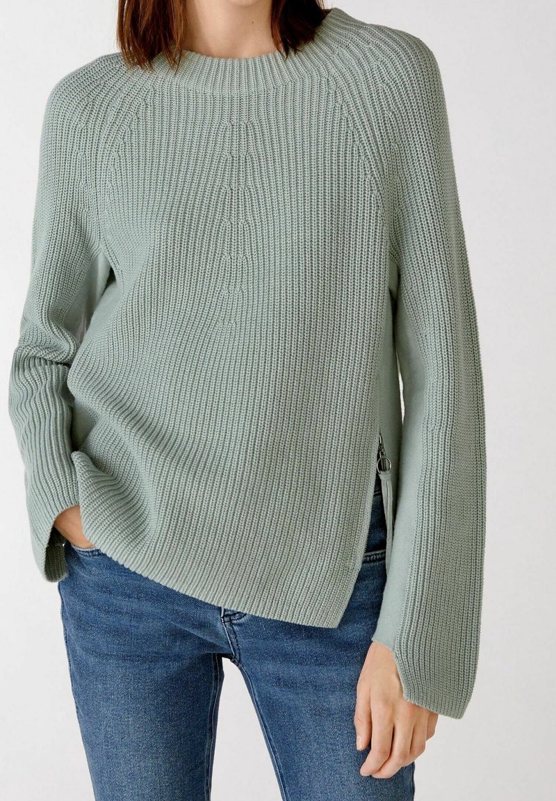 Light green knit sweater with ribbed texture, boat neckline, side slits, and long sleeves. Paired with blue denim jeans.