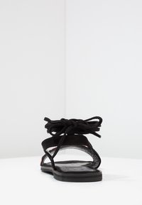 Black suede sandal with a flat sole, open toe design, and knotted strap detail at the ankle. Features soft texture and minimalistic style.