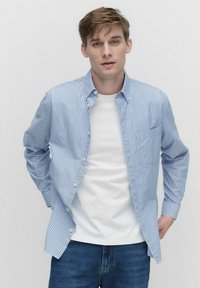 Blå og hvidstribet skjorte med knapper, blød krave, brystlomme og afslappet pasform over en hvid t-shirt; båret med denim jeans.