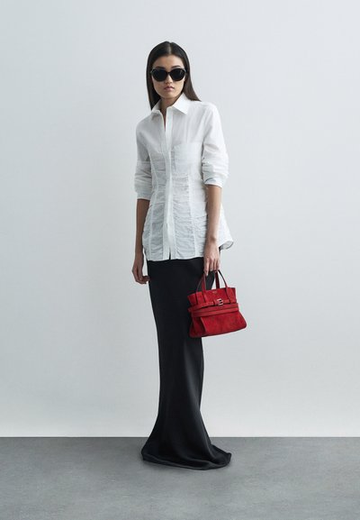 Kvinna som bär svarta solglasögon, vit knytblus, lång svart kjol, håller en handväska av röd mocka mot en enfärgad bakgrund.