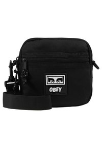 Czarna torba na ramię wykonana z trwałego materiału, z zamkiem błyskawicznym, regulowanym paskiem oraz przednią naszywką z logo "OBEY" i graficznymi detalami.