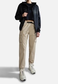 Osoba w beżowych spodniach cargo, białych trampkach, czarnej krótkiej bluzie i rozpiętej ciemnej kurtce z łańcuszkiem na szyi.