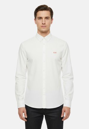 Uomo che indossa una camicia bianca aderente a maniche lunghe con bottoni e un piccolo logo rosso sul petto a sinistra, abbinata a pantaloni scuri.