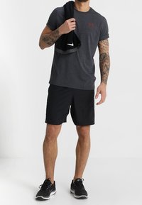 Tmavě šedé sportovní tričko s krátkým rukávem a červeným logem, černé kraťasy a černé běžecké boty. Model drží černý ručník.