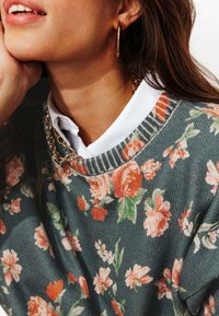 Pull à motif floral en bleu sarcelle avec des fleurs rouges et roses. Encolure côtelée, porté par-dessus une chemise blanche à col, avec des boucles d'oreilles hoops dorées.