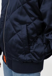 Navy gewatteerde bomberjack in glanzende stof met rib gebreide manchetten en zoom, voorzien van een ritsluiting en een zijzak. Details met oranje label zijn inbegrepen.