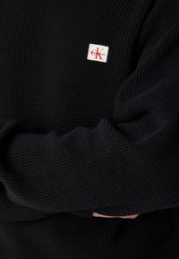 Zwart gebreid sweater met een textuur op het oppervlak en een klein rechthoekig Calvin Klein logo patch in rood en wit.