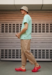 Rode klompen met een gestructureerd oppervlak, beige emmerhoed, lichtgroen T-shirt en beige opgerolde broek tegen een grijze achtergrond.