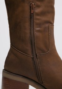 Botte de cheville en faux cuir marron avec fermeture éclair sur le côté, talon empilé et surpiqûres contrastantes le long de la couture pour un effet texturé.