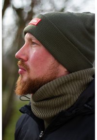 Olive green knit beanie with patch, paired with a textured olive green scarf and a black zip-up jacket, showcasing a close side profile.