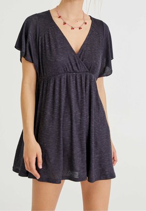 Donna con un vestito corto scuro a coste, maniche a sbuffo e una collana rosa di perline con piccoli fiori rossi.