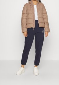 Giacca a vento color marrone chiaro con colletto alto, abbinata a pantaloni jogger blu navy con logo GAP. Il modello indossa scarpe da ginnastica bianche.