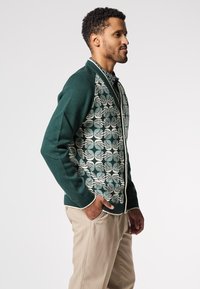 Pletený svetr ve tmavě zelené barvě s krémovým vzorem listu na přední straně. Žebrované manžety a lem s zapínáním na zip. Přiléhavý střih.