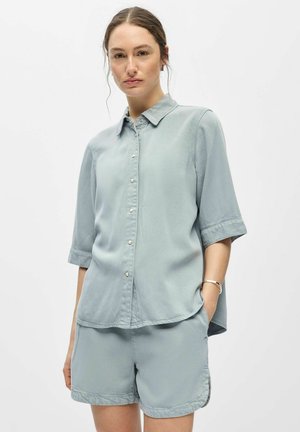 Femme portant une chemise bleu clair à boutons avec des manches jusqu'aux coudes et un short assorti, debout avec une main dans la poche.