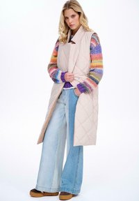 Kurtka bez rękawów, pikowana, w jasnoróżowym kolorze, noszona na multikolorowym, paskowanym swetrze. Szerokie, jasnoniebieskie dżinsy i brązowe buty na sznurówki dopełniają stylizację.