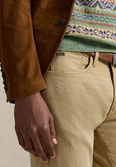 Blazer castanho em camurça, suéter multicolorido de malha, bainha canelada verde claro, calças cáqui com etiqueta de logótipo, cinto de couro castanho, tecido texturado.