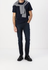 Navy t-shirt, grey sweater draped over shoulders, dark blue jeans, and black shoes. Simple design, smooth texture, and fitted silhouette.