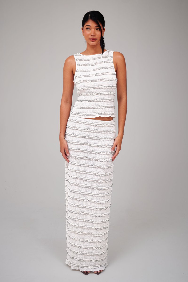 Vrouw met donker haar in een wit mouwloos topje en bijpassende lange rok met horizontale, gestructureerde ruches, staand tegen een effen grijze achtergrond.