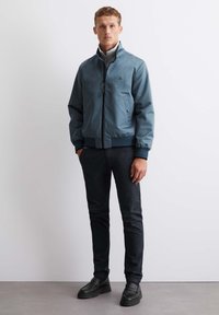 Blue bomber jacket with a zip front, ribbed cuffs, and two side pockets, worn over a gray high-collar sweater and paired with black pants.