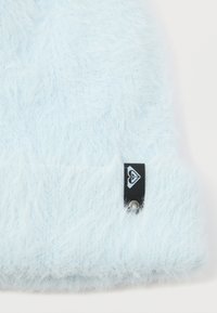 Gorro de malha fofinho, azul claro, com um acabamento texturizado. Apresenta uma etiqueta preta com um logótipo e um detalhe em metal, acrescentando pormenor ao design.