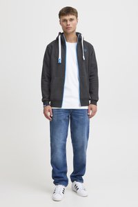 Haut de survêtement zippé gris foncé avec des accents bleus, t-shirt blanc, jeans bleus et baskets blanches avec des rayures noires. Tissu doux, coupe décontractée.