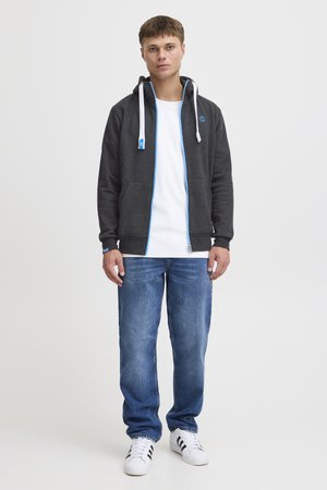 Ciemnoszary hoodie zapinany na zamek z niebieskimi akcentami, biały t-shirt, niebieskie dżinsy i białe sneakersy z czarnymi paskami. Miękki materiał, luźny krój.