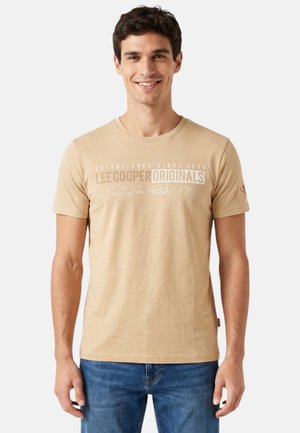 Jeune homme souriant, portant un t-shirt beige Lee Cooper Originals et un jean bleu, debout devant un fond clair uni.