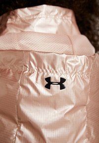 Kurtka pikowana w jasnoróżowym kolorze, o gładkiej, błyszczącej fakturze z żeberkowanymi elementami wzorniczymi. Na kurtce widoczne jest czarne logo Under Armour.