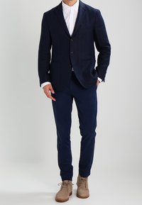 Man klädd i marinblå kostym med vit skjorta och beige mockaskor med snörning, står mot en enfärgad bakgrund.