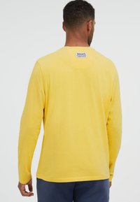 Mann trägt ein gelbes Langarmshirt mit einem "No #1 focus on colour CAMP DAVID"-Label auf dem oberen Rücken, kombiniert mit dunkler Hose.