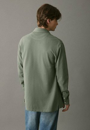 Zelená košeľa s dlhým rukávom a golierom, s voľným strihom a gombíkovými manžetami, v kombinácii so svetlo modrými džínami. Hladký materiál.
