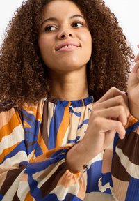 Mehrfarbige Bluse mit blauen, orangen, braunen und cremefarbenen abstrakten Mustern. Lange, gepuffte Ärmel mit gerafften Manschetten und einem runden Halsausschnitt.