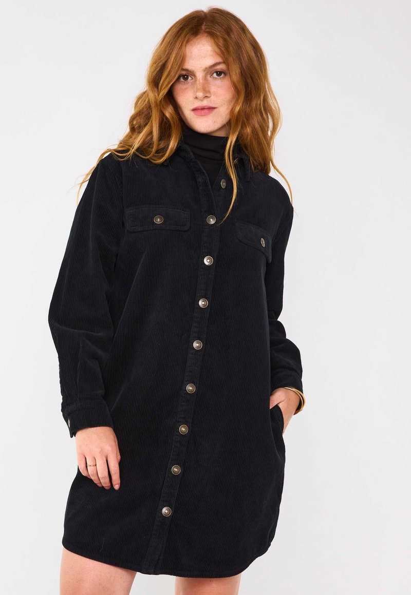 Abito camicia in corduroy di colore blu navy scuro, con maniche lunghe, chiusura frontale a bottoni, taschini sul petto e tasche laterali. Presenta dettagli in metallo sui bottoni.