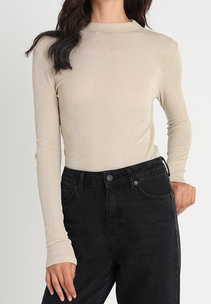 Femme portant un haut beige ajusté à manches longues rentré dans un jean noir taille haute, debout avec une main posée sur la hanche.