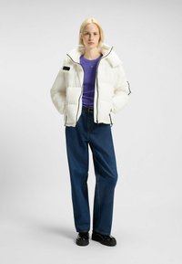 Vit puff-jacka med stora kviltade sektioner, svarta dragkedjor och hög krage. Tillsammans med en lila skjorta och vida jeans.