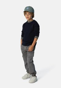 Suéter navy, pantalones cargo grises con bolsillos y zapatillas blancas; gorra verde con detalle de logo, puños en capas para textura y contraste.