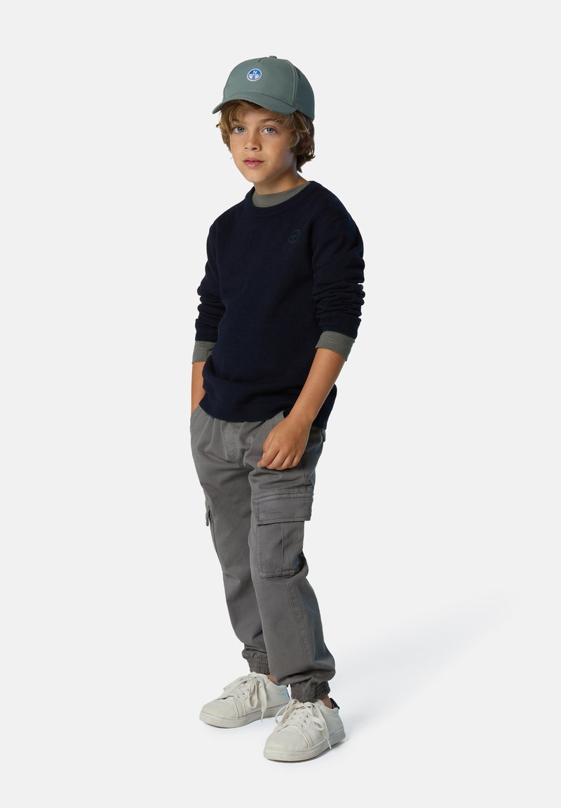 Suéter navy, pantalones cargo grises con bolsillos y zapatillas blancas; gorra verde con detalle de logo, puños en capas para textura y contraste.