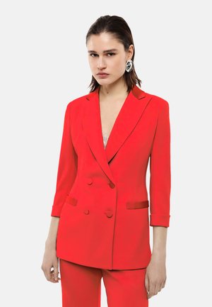 Donna che indossa un blazer doppiopetto rosso brillante con pantaloni abbinati e grandi orecchini a cerchio argentati, in piedi davanti a uno sfondo semplice.