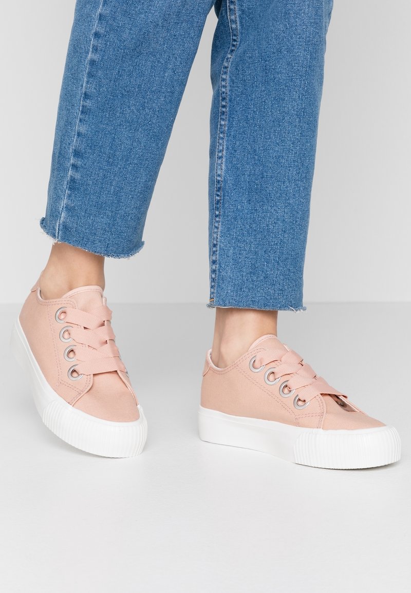 Osoba ubrana w niebieskie krótkie dżinsy i jasnoróżowe płócienne trampki z grubymi białymi gumowymi podeszwami i szerokimi płaskimi sznurówkami na szarym podłodze.