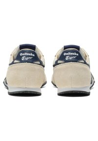 Sneakers in suede beige con accenti blu navy, dotate di un logo sul tallone e una suola texturizzata per una buona aderenza. Design classico con dettagli minimi.