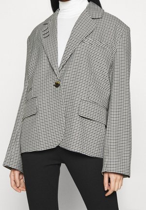 Femme portant un blazer pied-de-poule noir et blanc à un bouton, sur un col roulé blanc et un pantalon noir.