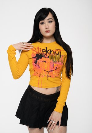 Yellow long-sleeve crop top featuring black and pink graphic print with drips, paired with a black pleated mini skirt.